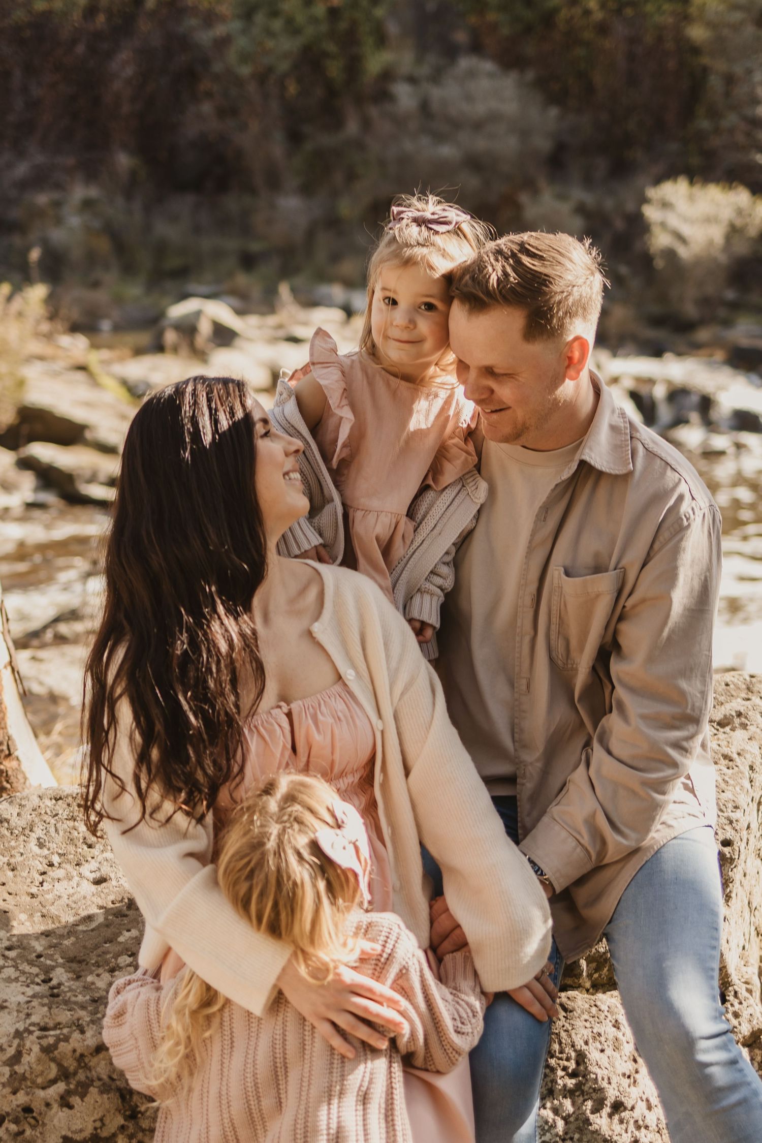 A happy family of four poses outdoors in warm neutral tones, with parents embracing two young daughters in a sunny natural setting.