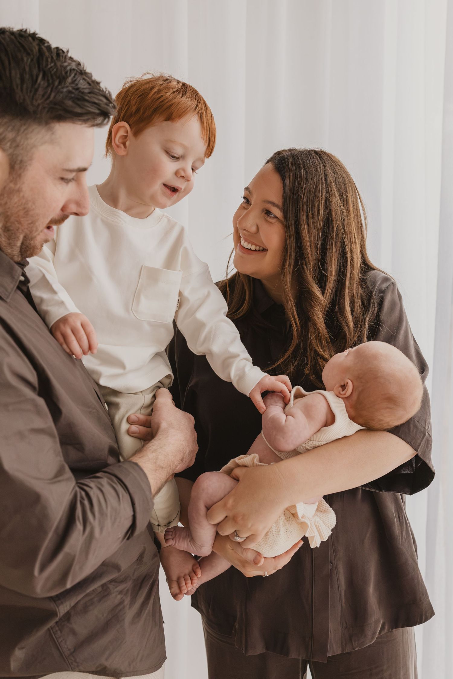 Happy family with parents holding a red-haired toddler and newborn baby, smiling together in a bright room.
