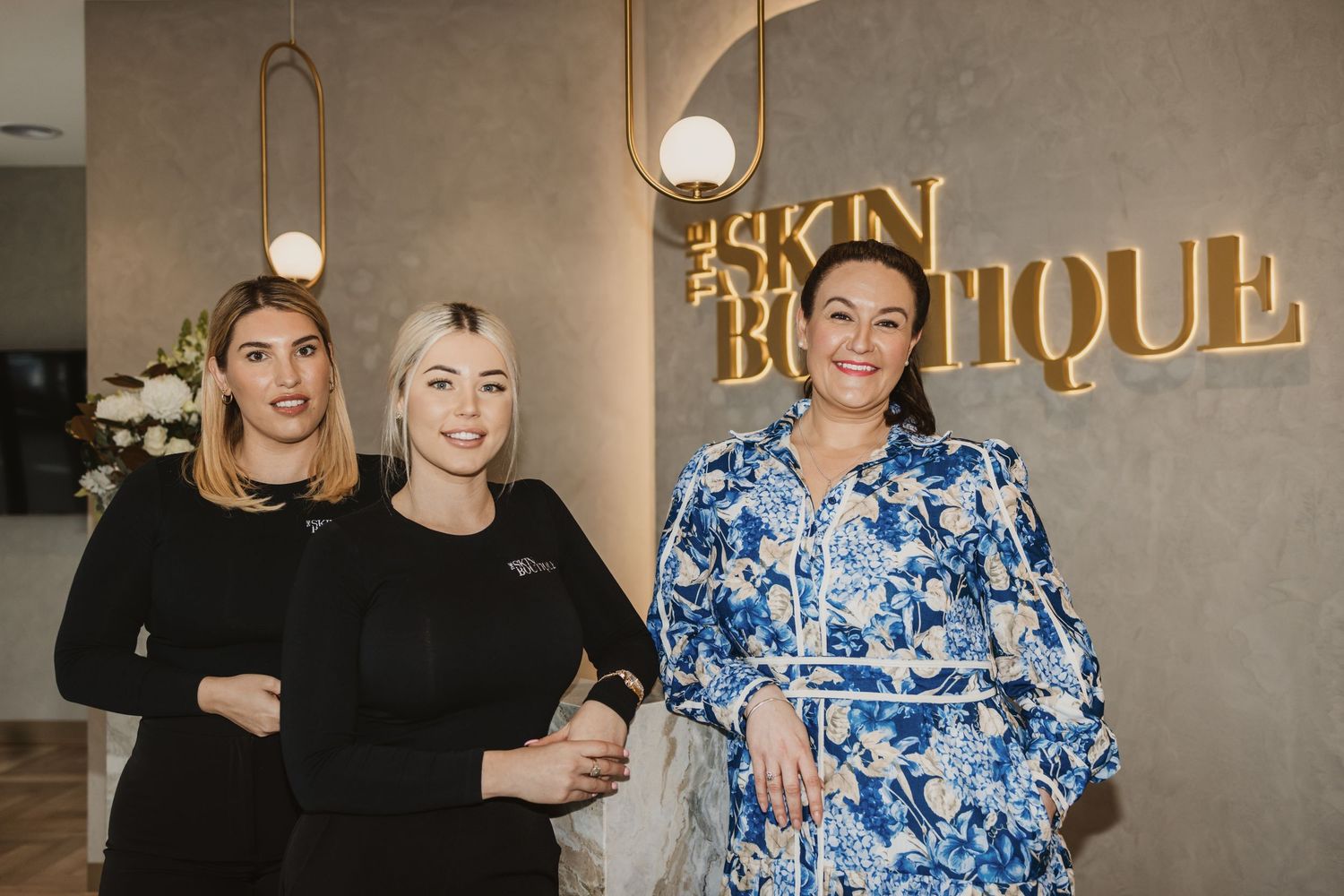 Three women posing together in front of The Skin Boutique salon sign, two in black uniforms and one in a floral dress.