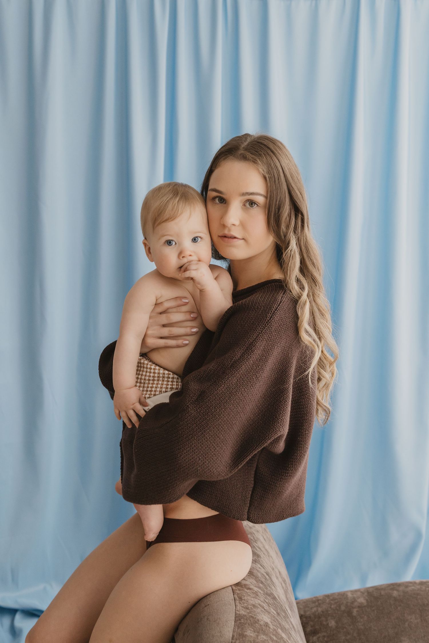 Young mother holding baby infant in brown sweater against blue curtain backdrop in studio portrait.