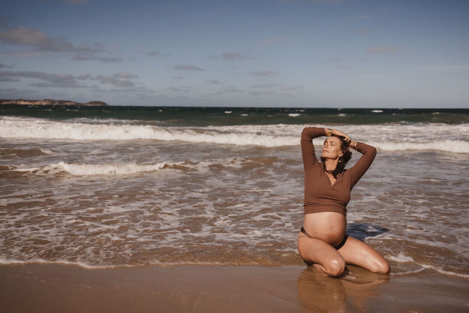 Pregnant woman in brown swimsuit kneeling on sandy beach with ocean waves, maternity photography outdoors.