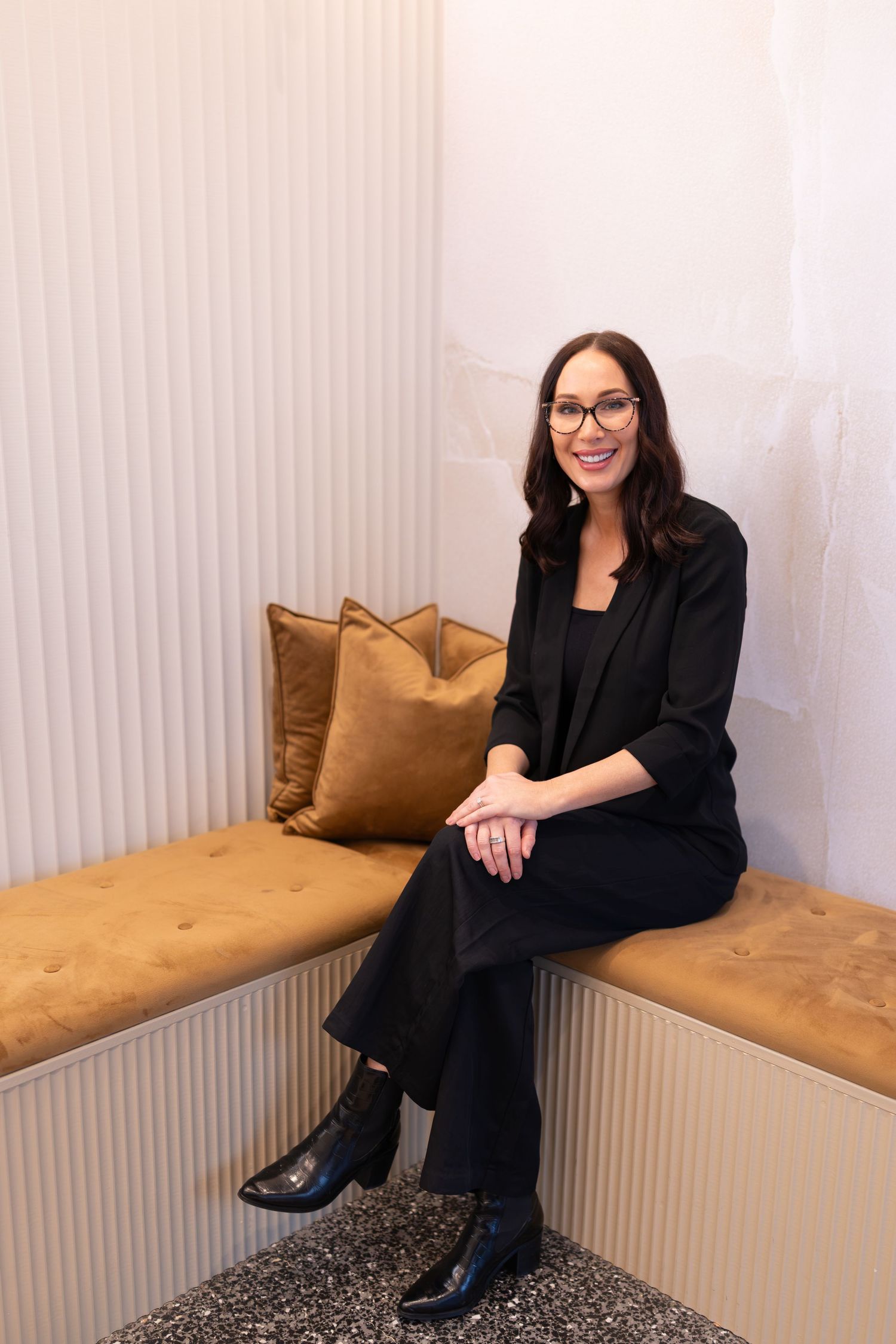 Woman in black blazer and pants sitting on a tan tufted bench, smiling, wearing glasses in a modern interior.