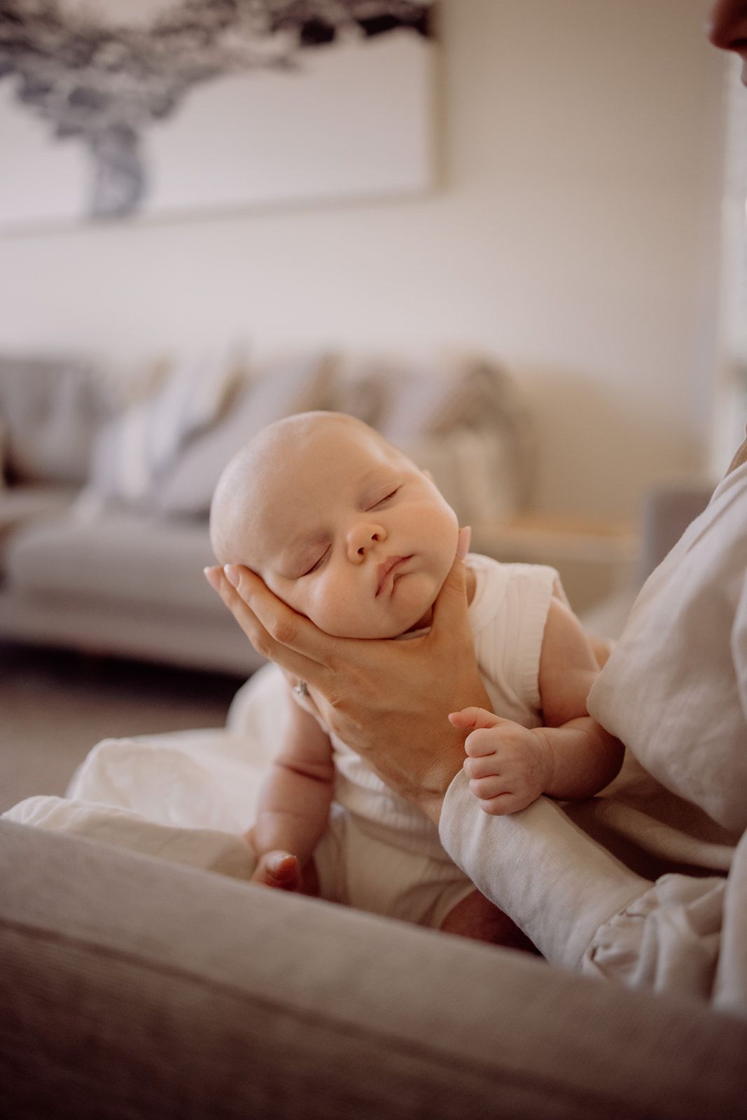 A newborn baby sleeping peacefully while being cradled in an adult's hand in a cozy living room.