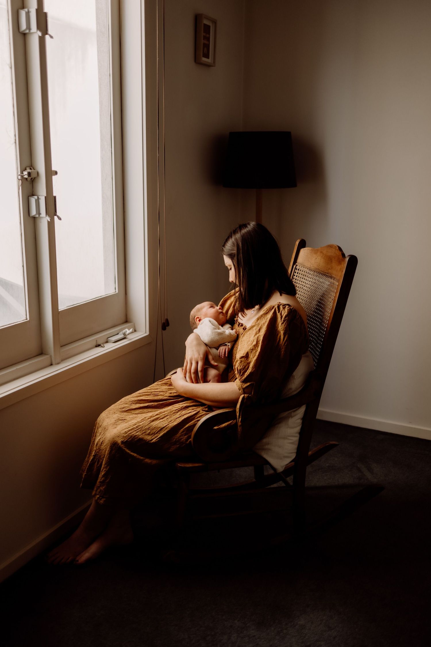 Mother in mustard dress sits in wooden chair holding newborn baby by window in warm natural light.