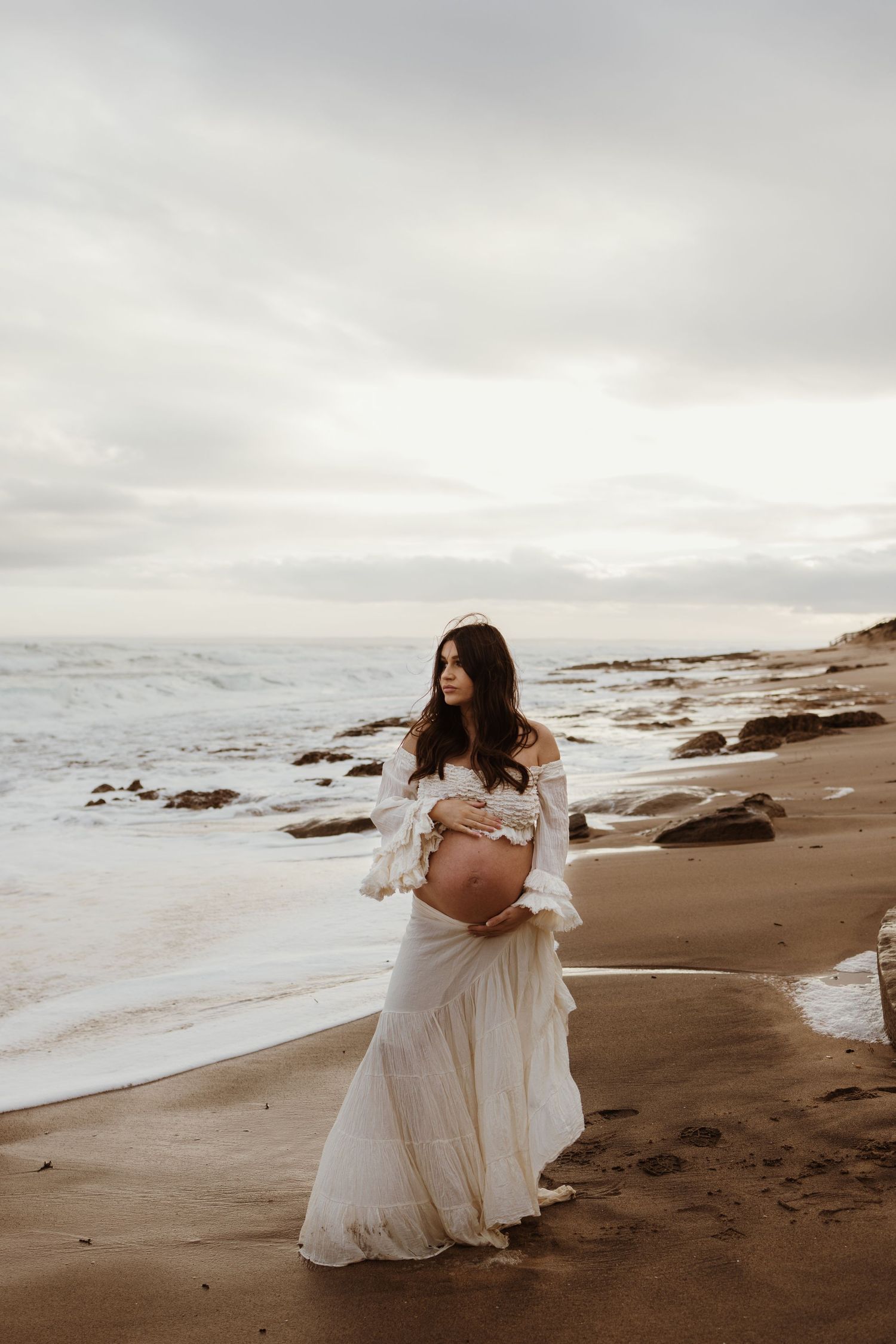 Pregnant woman in white boho dress holding baby bump on sandy beach with ocean waves and cloudy sky.