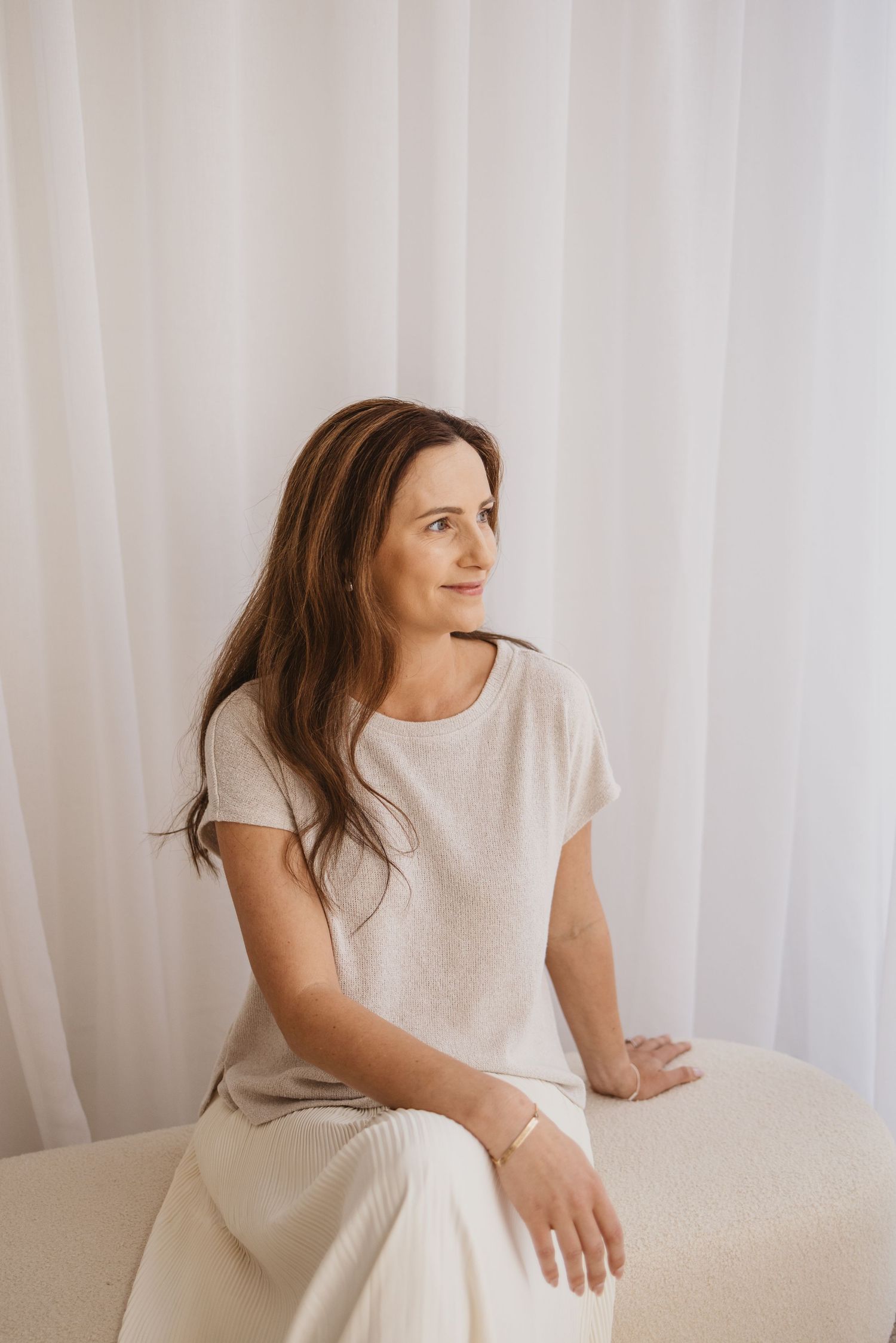 Woman in white outfit sitting peacefully against sheer curtains, gazing thoughtfully to the side in a bright minimalist space.