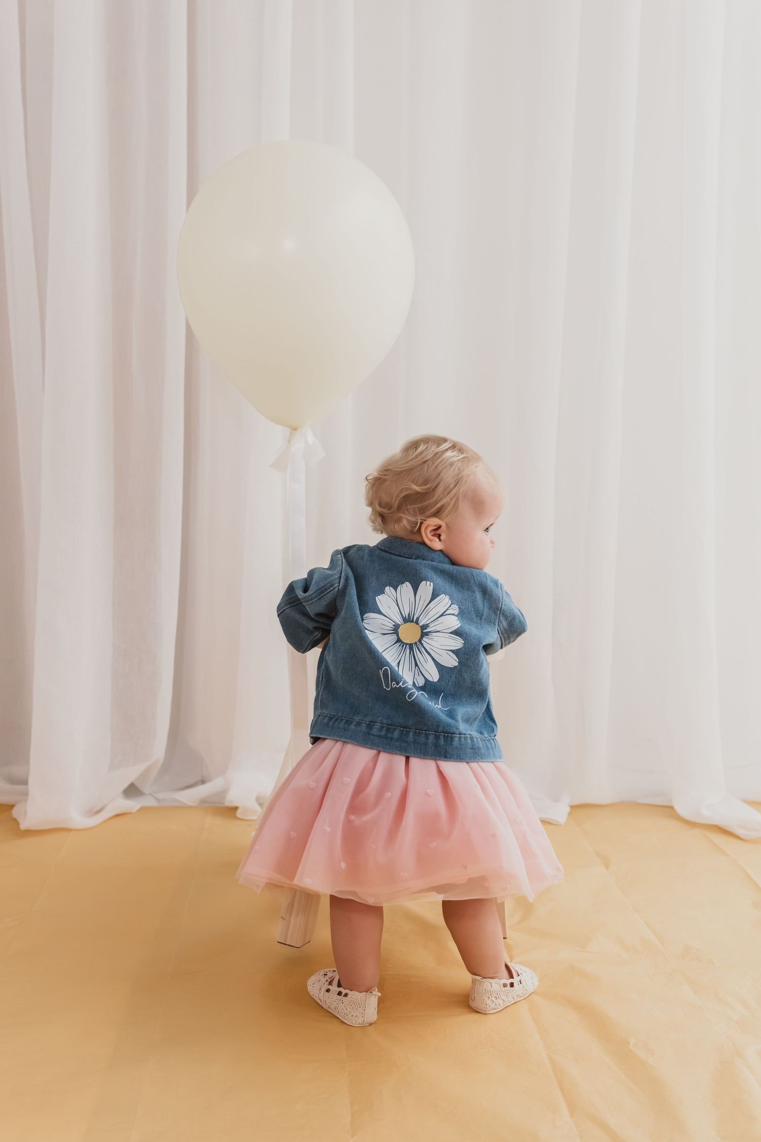 Toddler girl in denim daisy jacket and pink tutu skirt holding white balloon, viewed from behind.