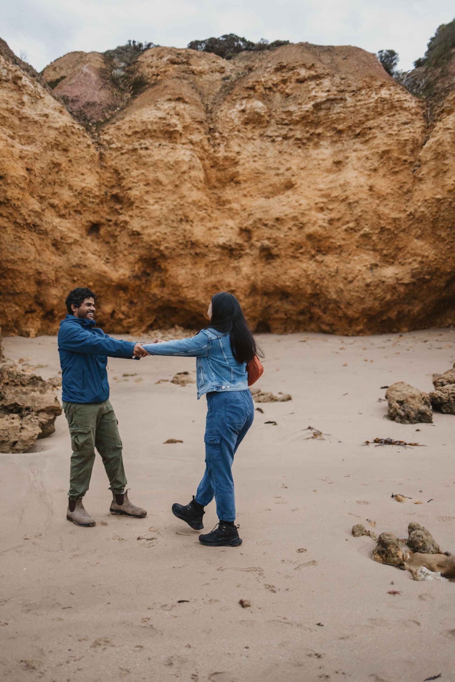 Couple holding hands and dancing on a sandy beach with dramatic golden rock cliffs in the background.