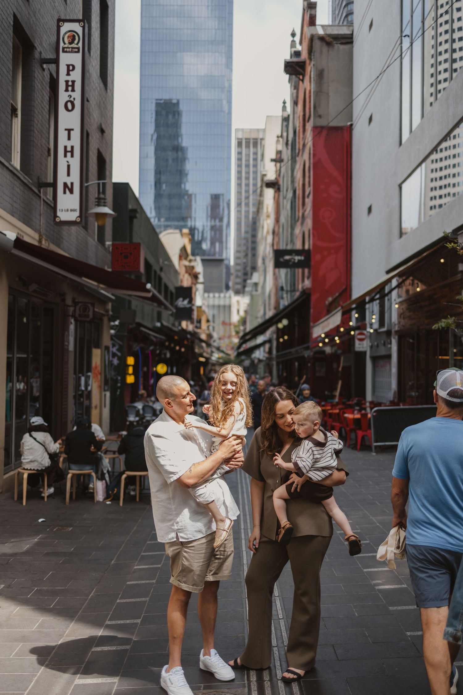 Family posing with two babies in Melbourne laneway near Pho Thin restaurant, city skyscrapers visible in background.