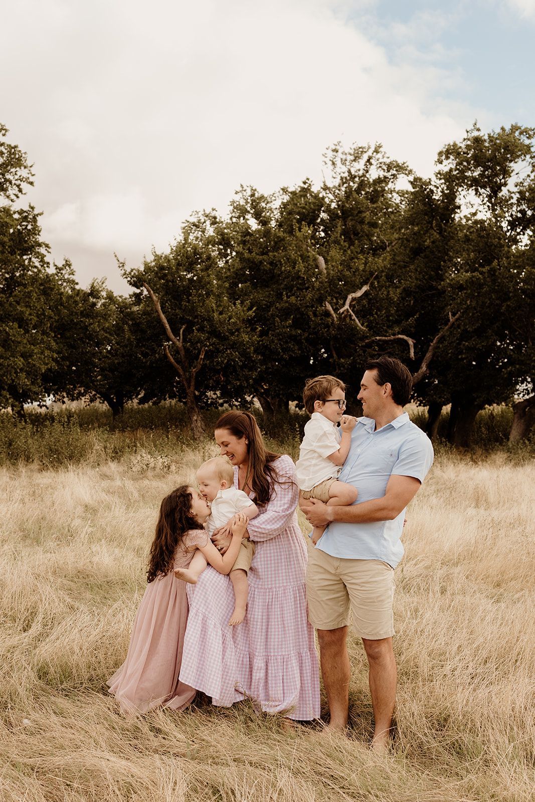 Family of four poses in golden field with trees, parents holding two young children, wearing pastel outfits on cloudy day.