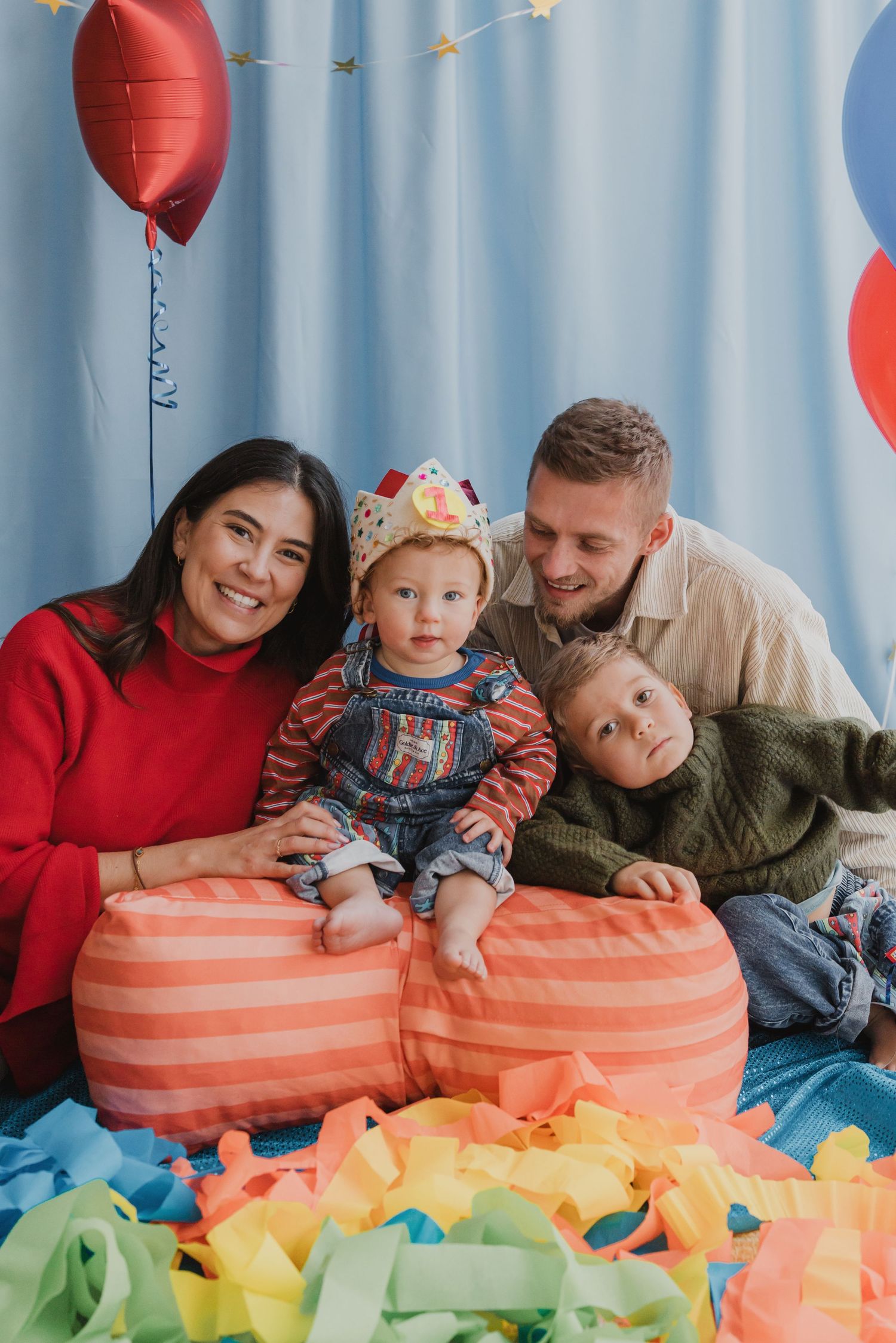 Happy family with two young children celebrating a birthday with colorful balloons and confetti indoors.