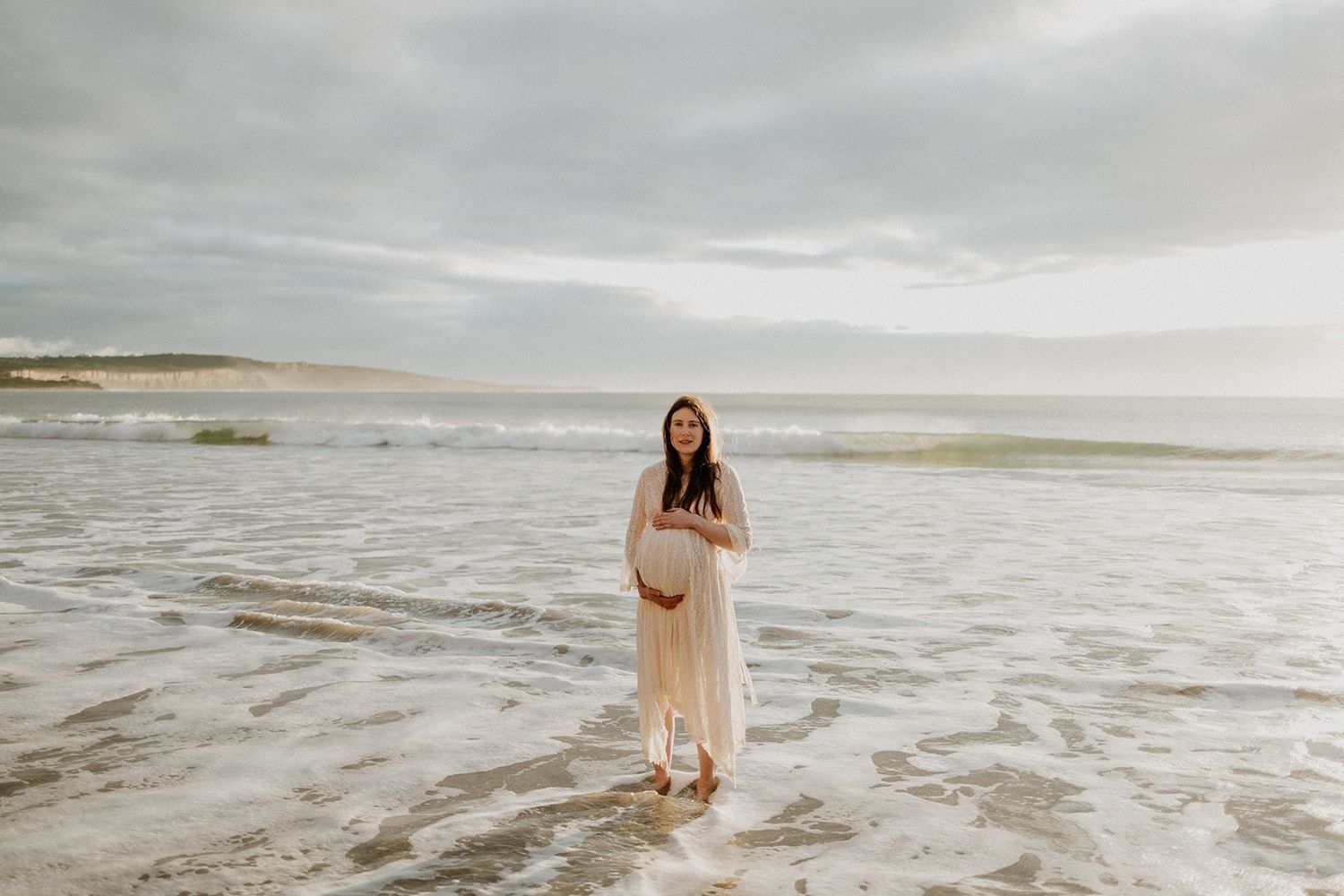 Pregnant woman in white dress standing barefoot on a serene beach with calm ocean waves and cloudy sky.