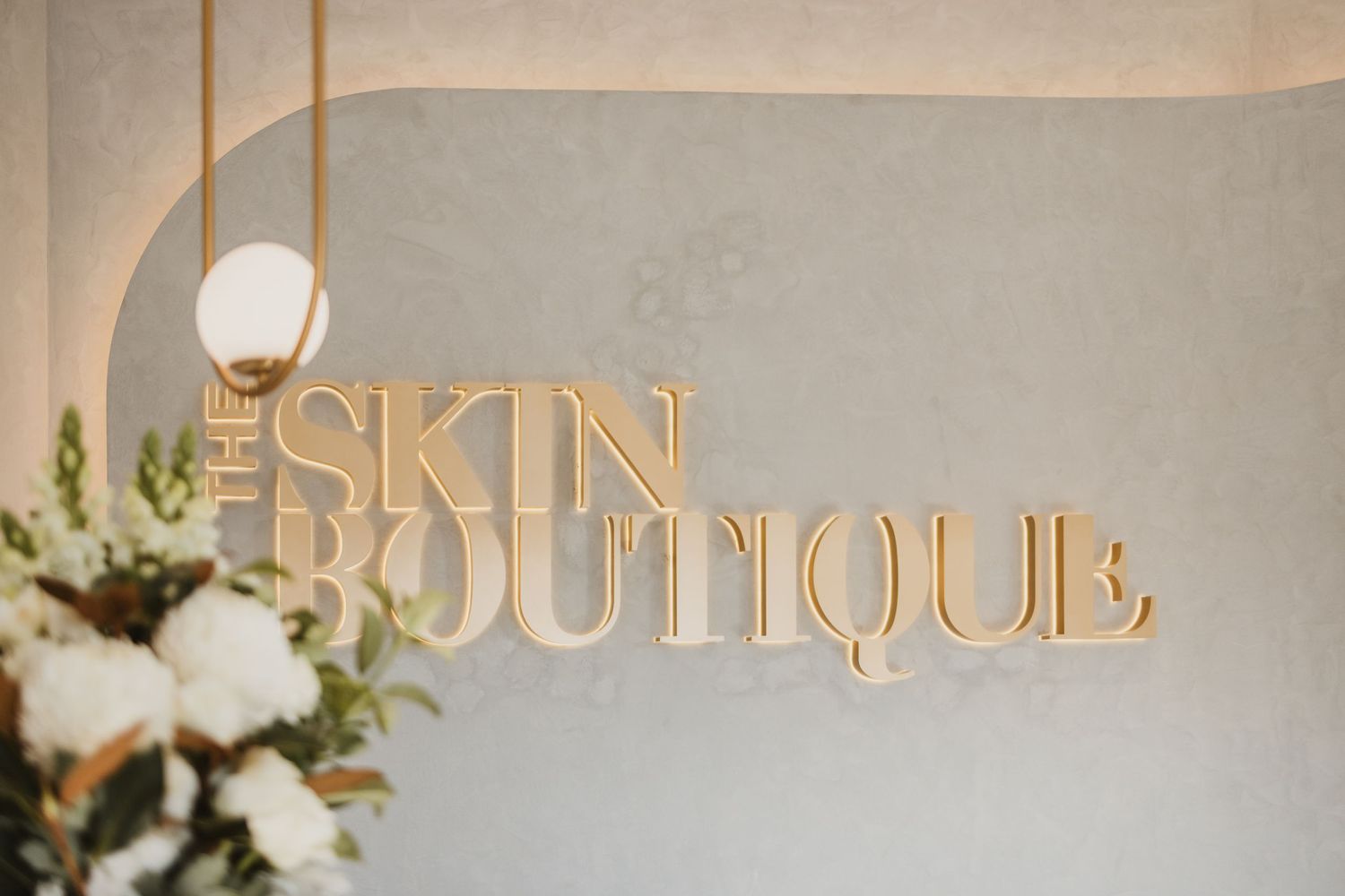 Gold 3D lettering of The Skin Boutique logo on a gray wall with a globe pendant light and white flowers.