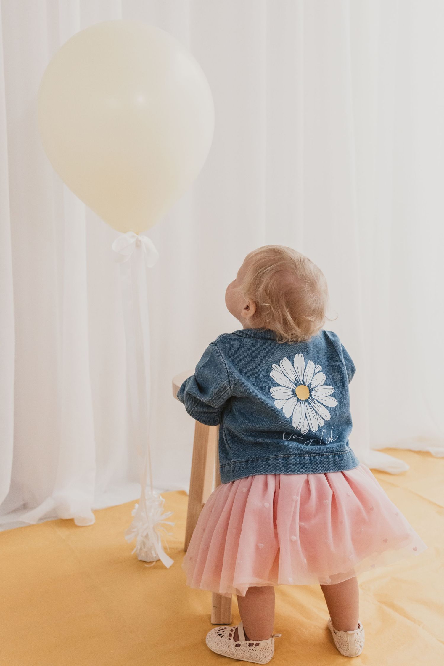 Toddler girl in denim daisy jacket and pink tutu skirt stands facing a white balloon near sheer curtains.