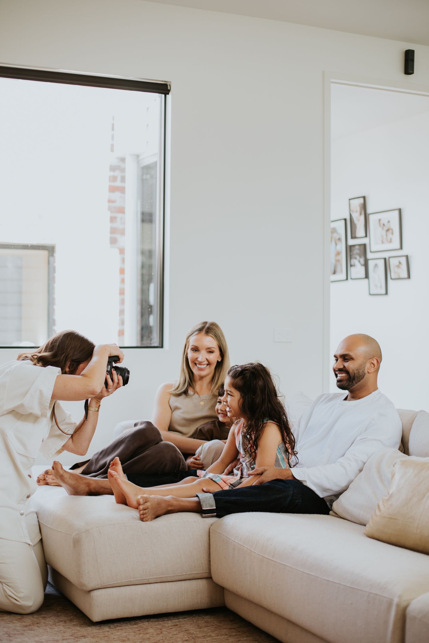Photographer capturing a happy family of three posing on a white sectional sofa in a bright modern living room.