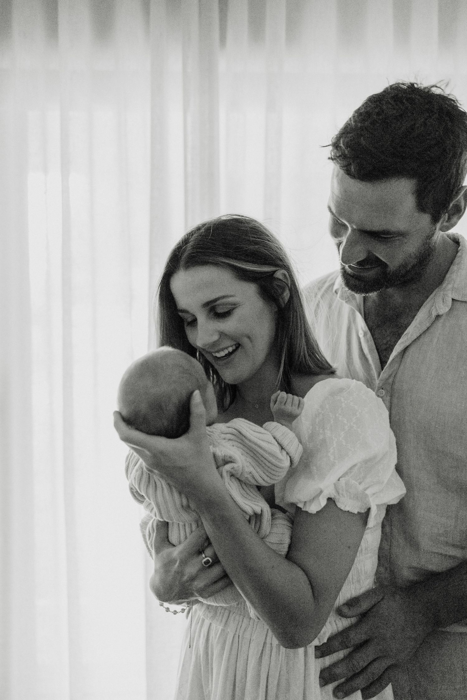 Black and white photo of smiling parents lovingly gazing at their newborn baby wrapped in white.
