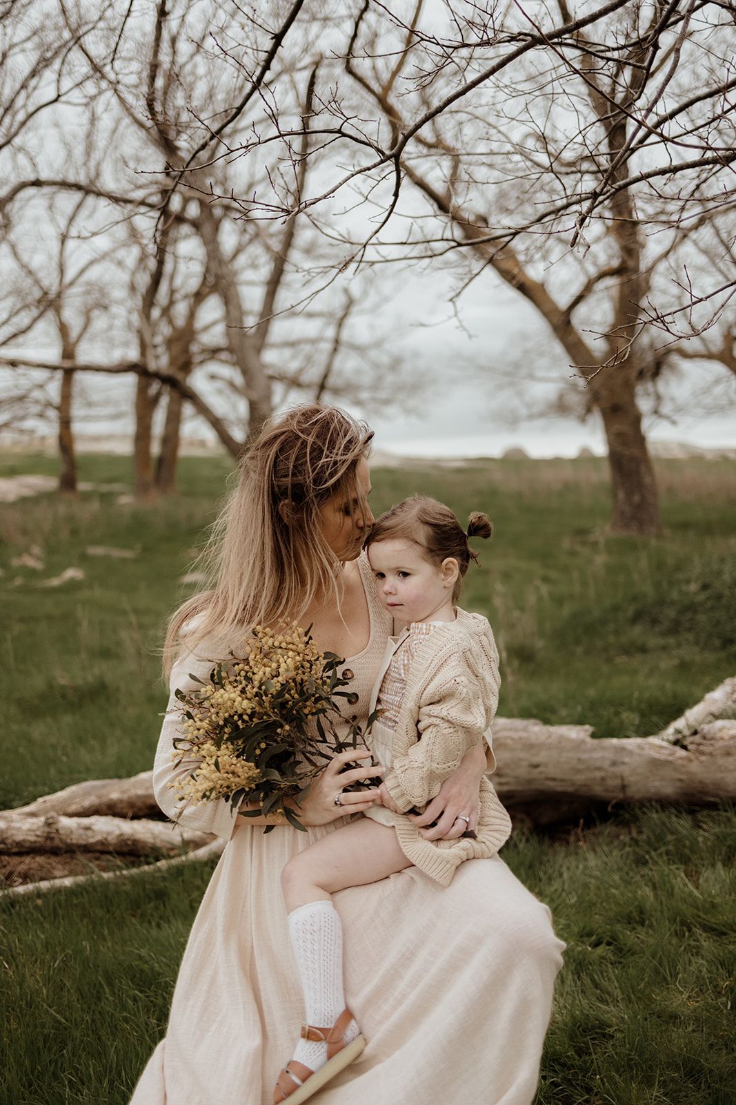 Mother in beige dress holds toddler girl with yellow wildflowers while sitting on fallen log among bare trees outdoors.