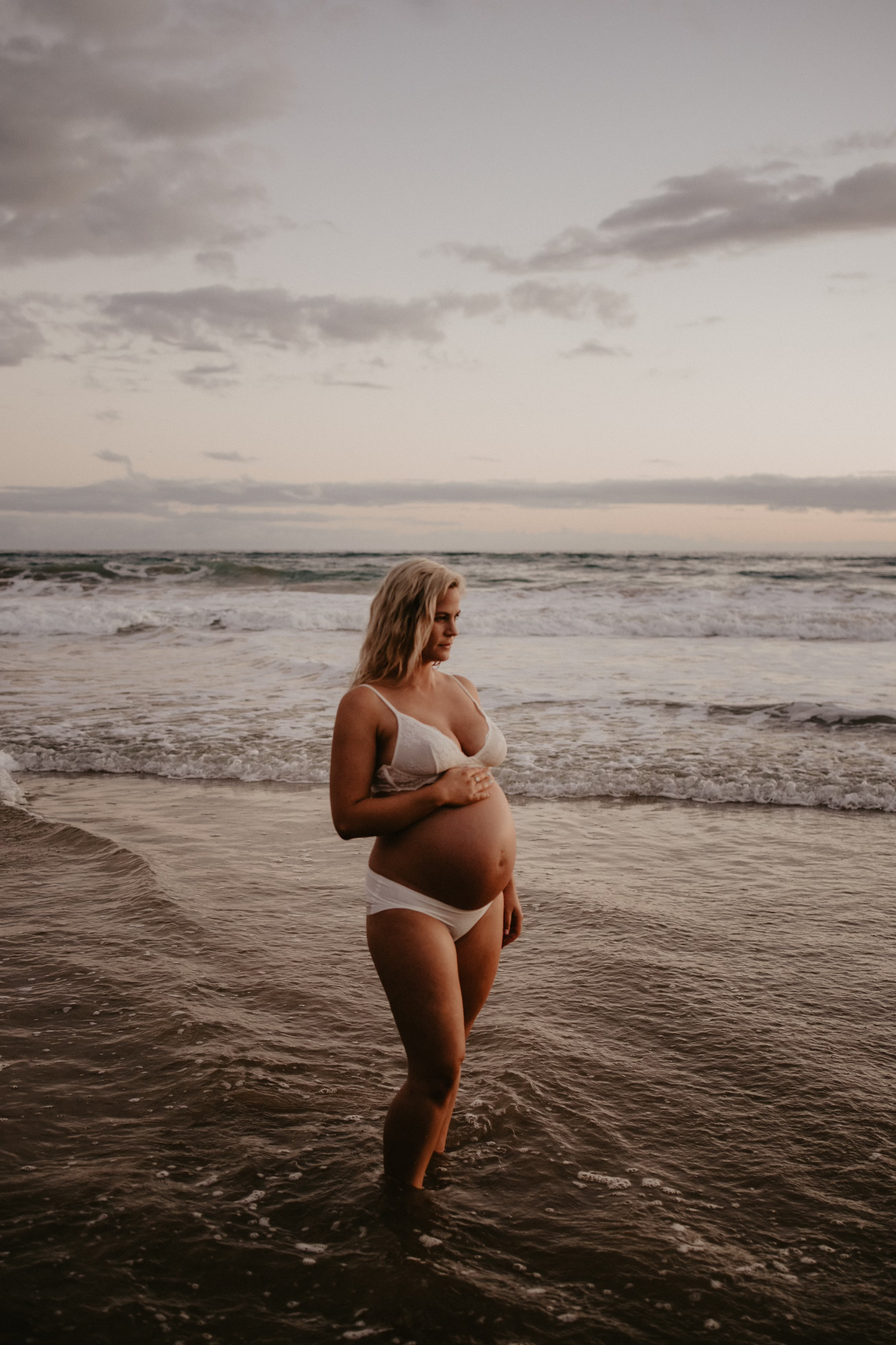 Geelong maternity photography - pregnant woman in white swimwear standing in the ocean at sunset, cradling her bump.