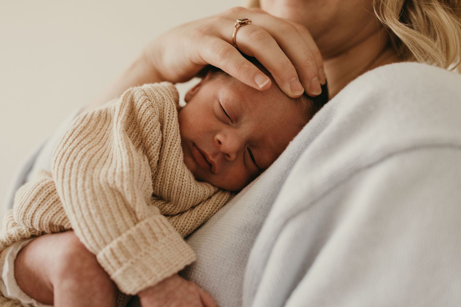 Sleeping newborn baby wearing a cream colored knit sweater rests peacefully against soft white fabric.