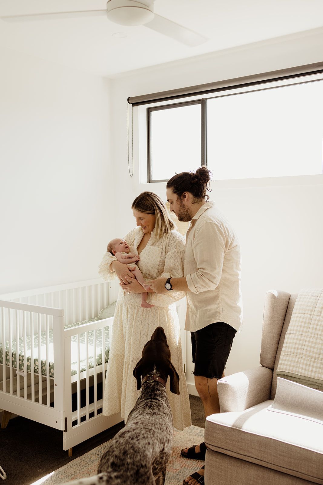 New parents with newborn baby and dog in bright nursery room with white crib and ceiling fan.