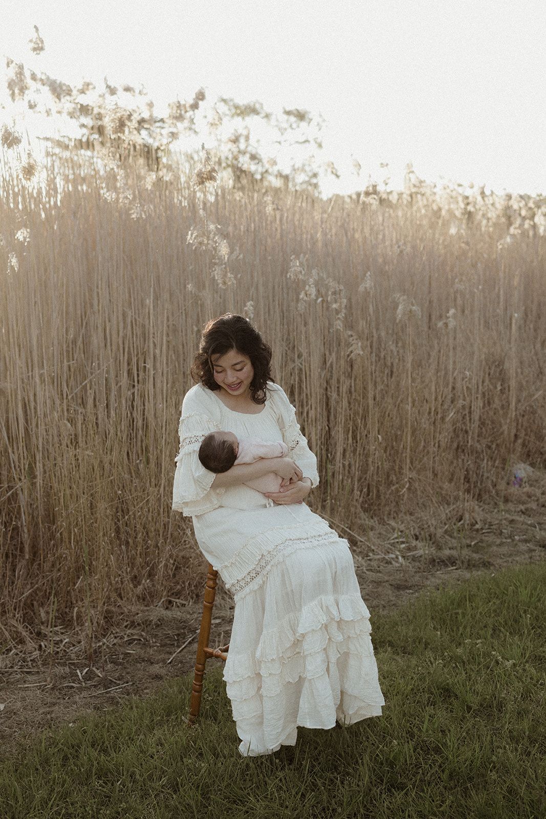 A mother in a white boho dress sits outdoors breastfeeding her newborn baby, surrounded by golden tall grass.