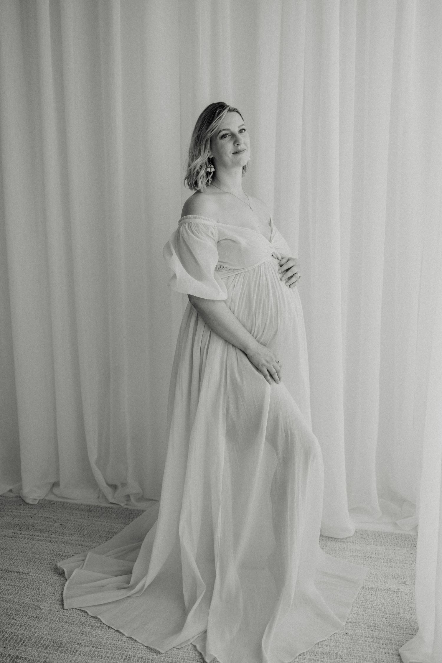 Pregnant woman in elegant white off-shoulder gown poses gracefully in black and white maternity portrait.