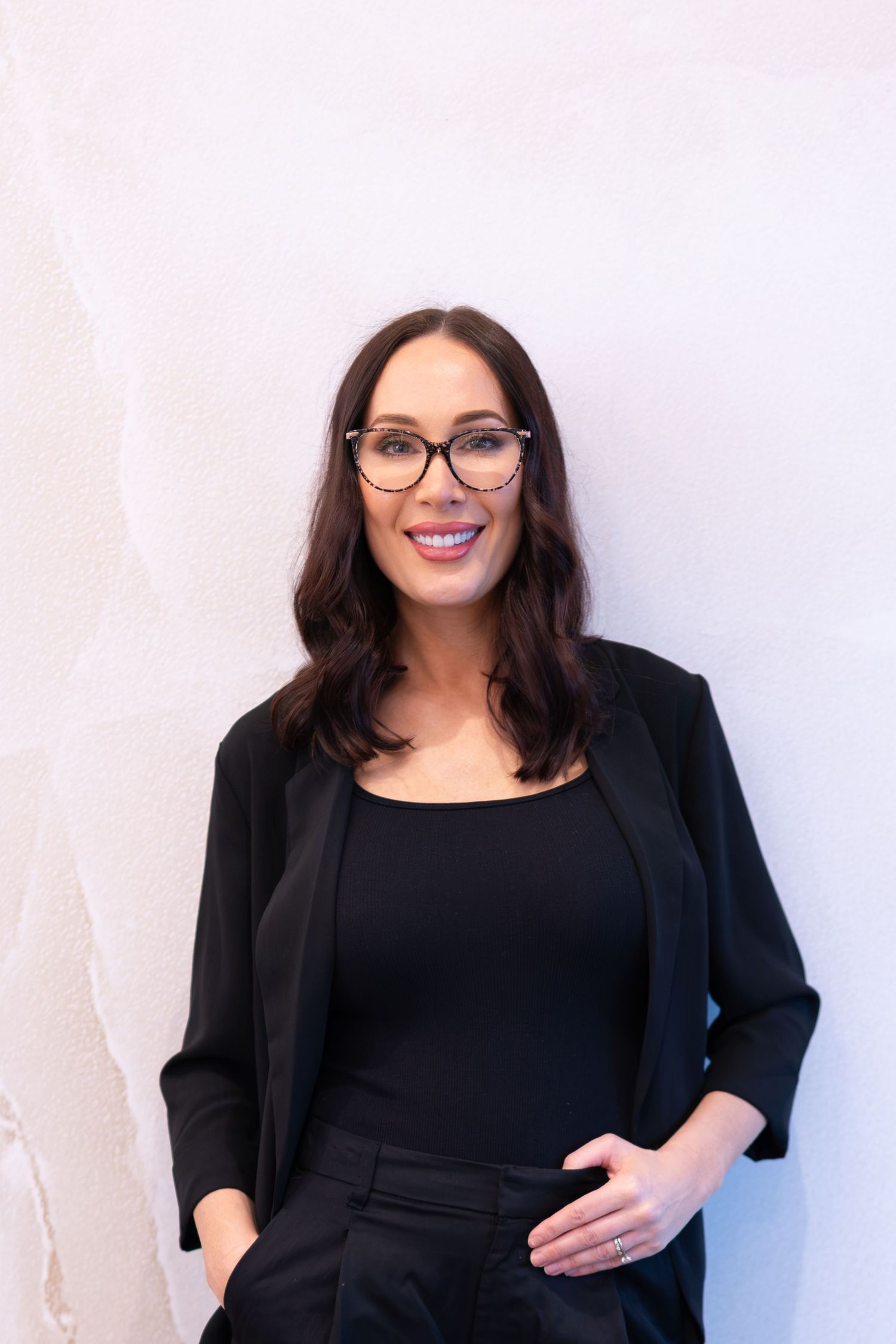 A smiling woman with dark hair wearing black glasses, a black blazer, and black top poses against a white wall.