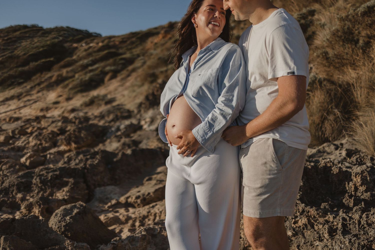 Pregnant woman in white outfit smiling with partner near rocky coastal cliffs during maternity photoshoot.