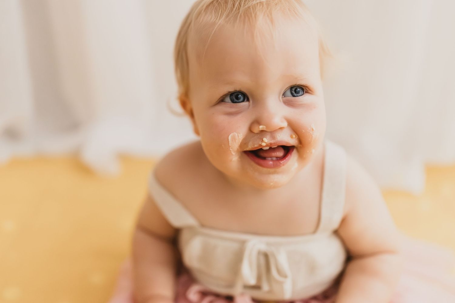 Adorable baby girl smiling with food on her face, wearing a linen romper during a cake smash photo session.