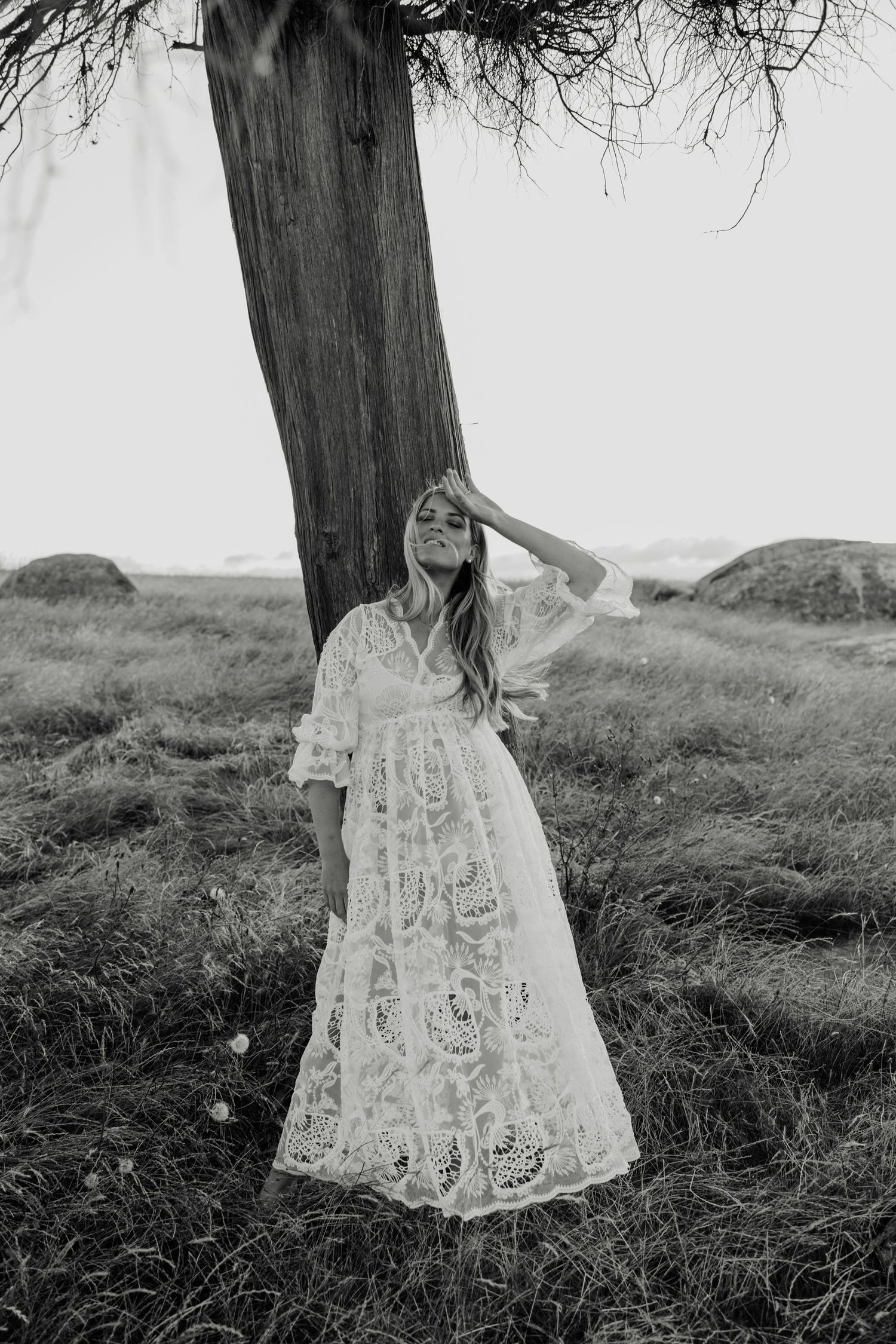 Woman in white lace maxi dress standing by a tree in a field, black and white photography.