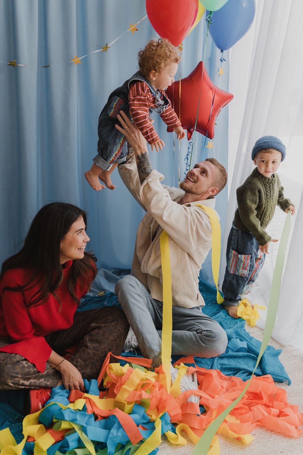 A joyful family celebrates a birthday with colorful balloons and streamers, dad lifting toddler while mom and child watch.