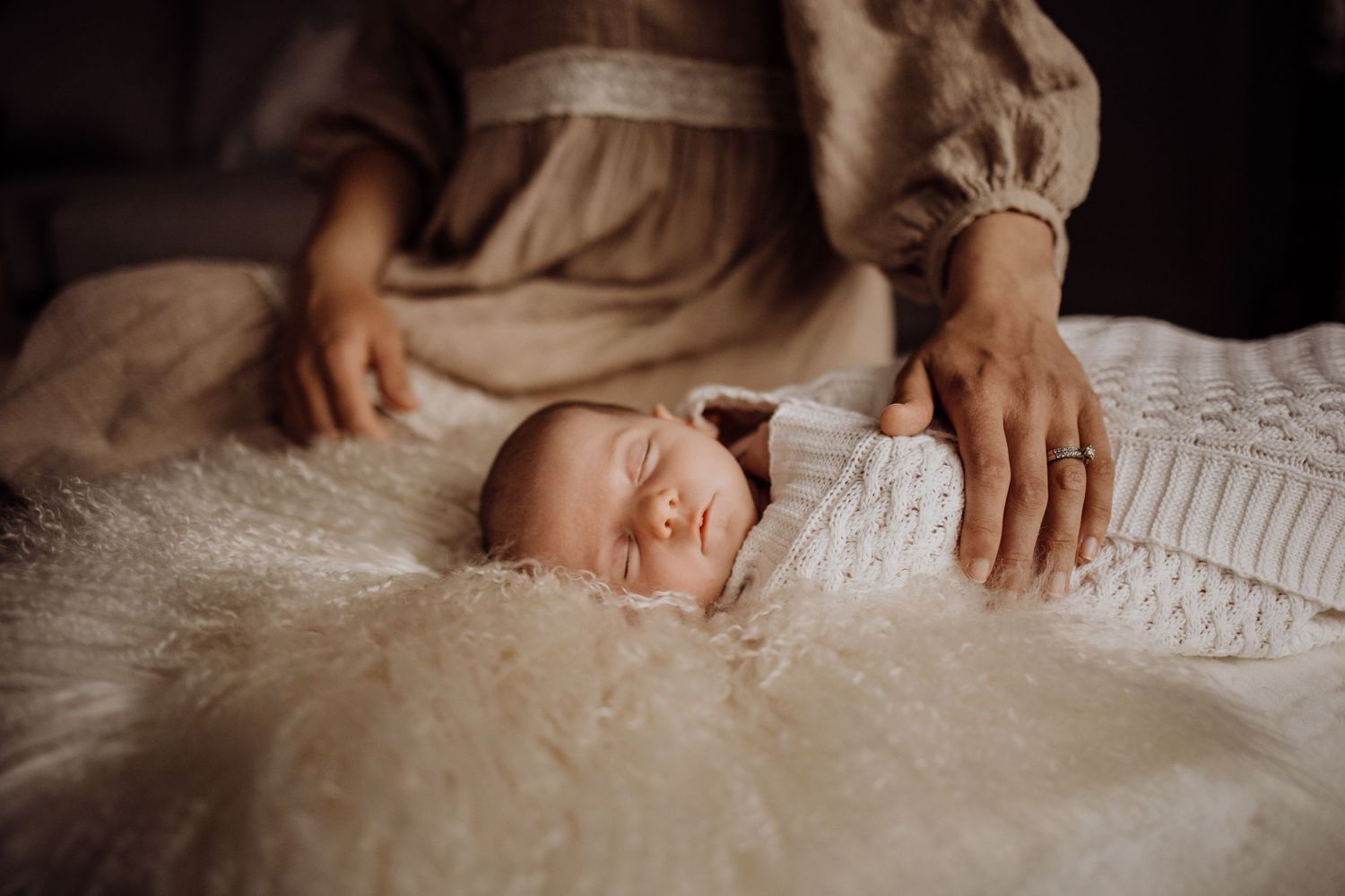 Newborn baby sleeping peacefully wrapped in white swaddle on fluffy blanket with mother in beige dress nearby.