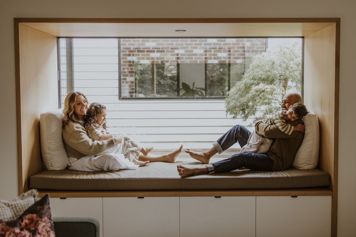 Two families relax together on a built-in window seat bench in a modern home with large windows.