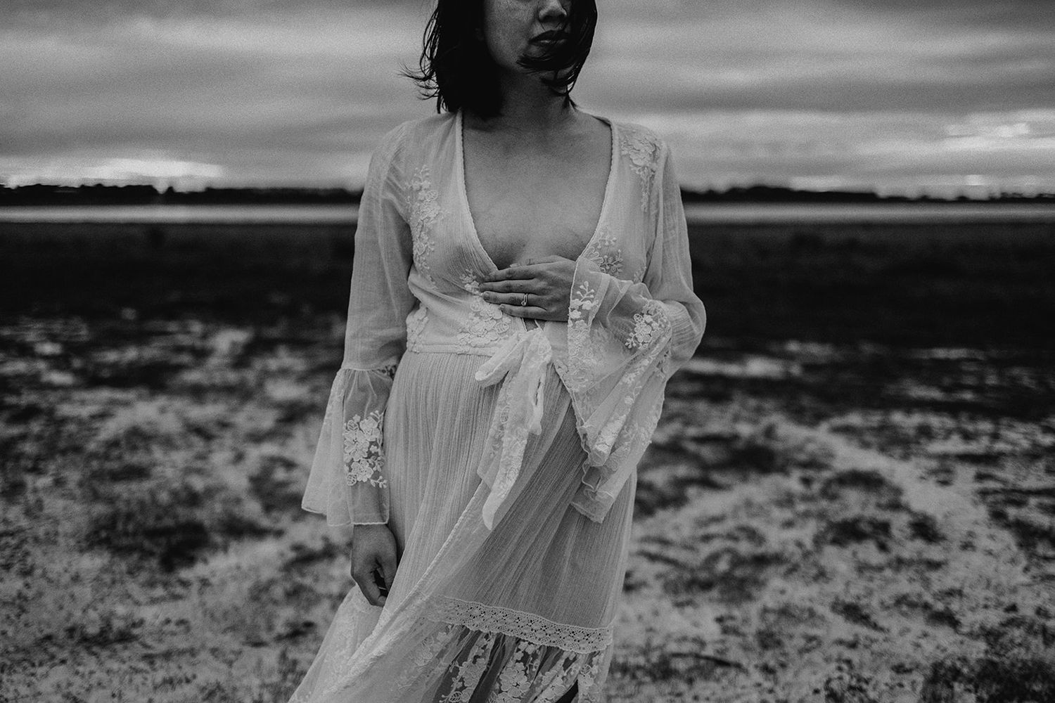 Black and white photo of a woman in a flowing white lace dress standing in an open field under a cloudy sky.