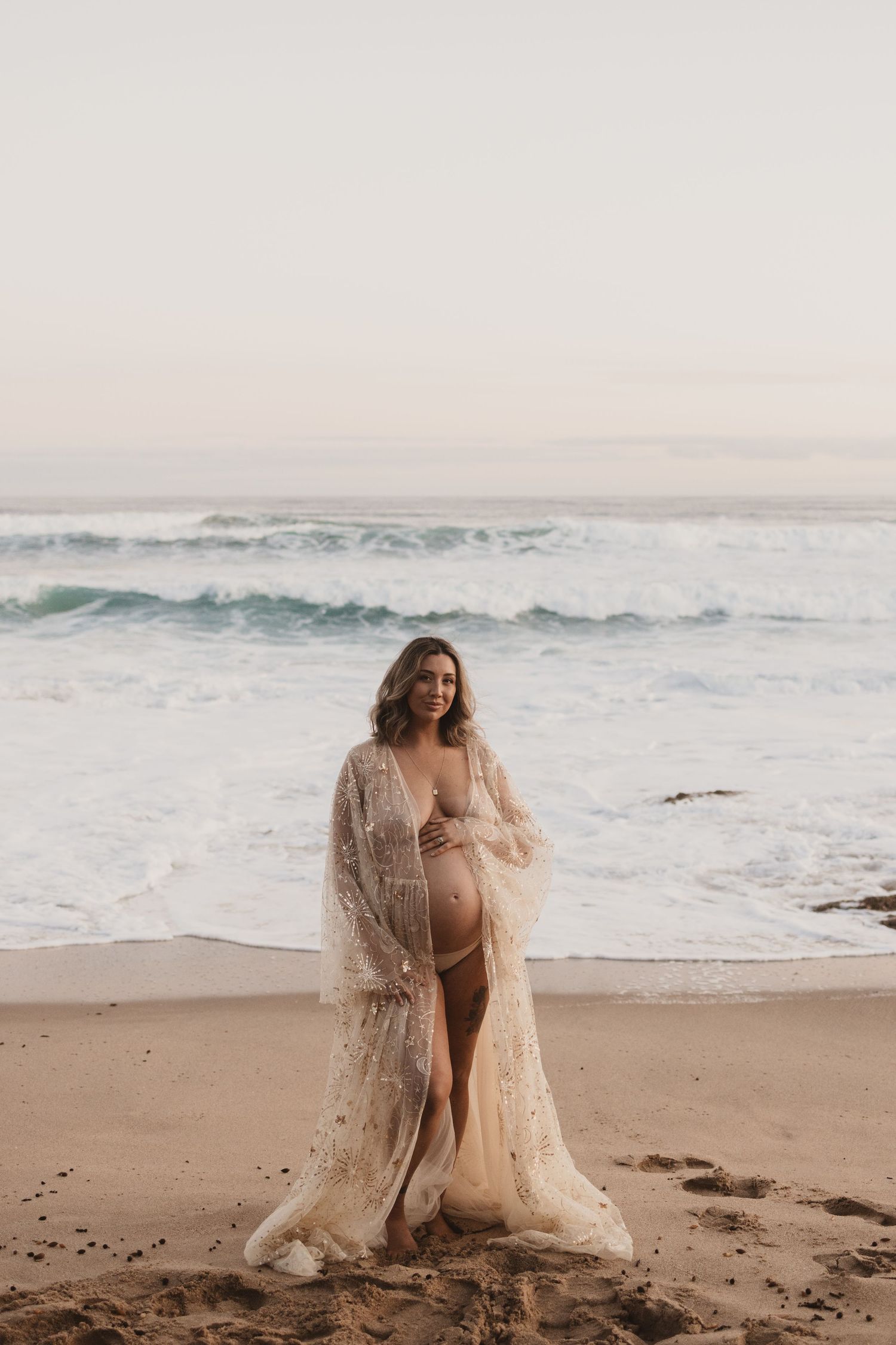 Pregnant woman in sheer lace robe poses on sandy beach with ocean waves, maternity photography.