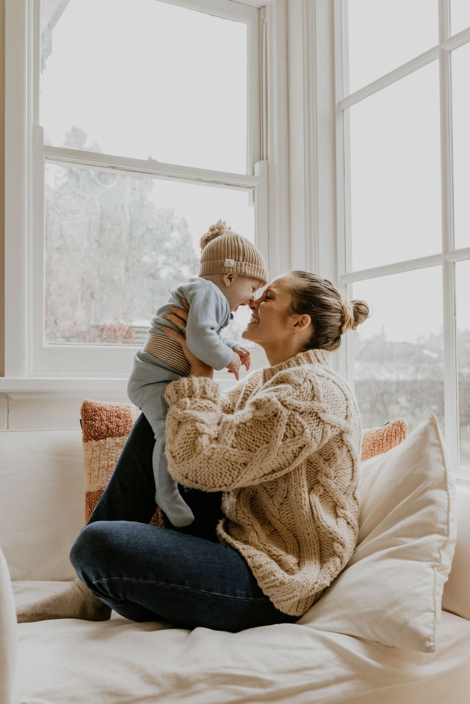 A mother in a cream knit sweater holds her baby in a beanie near a bright window on a white couch.