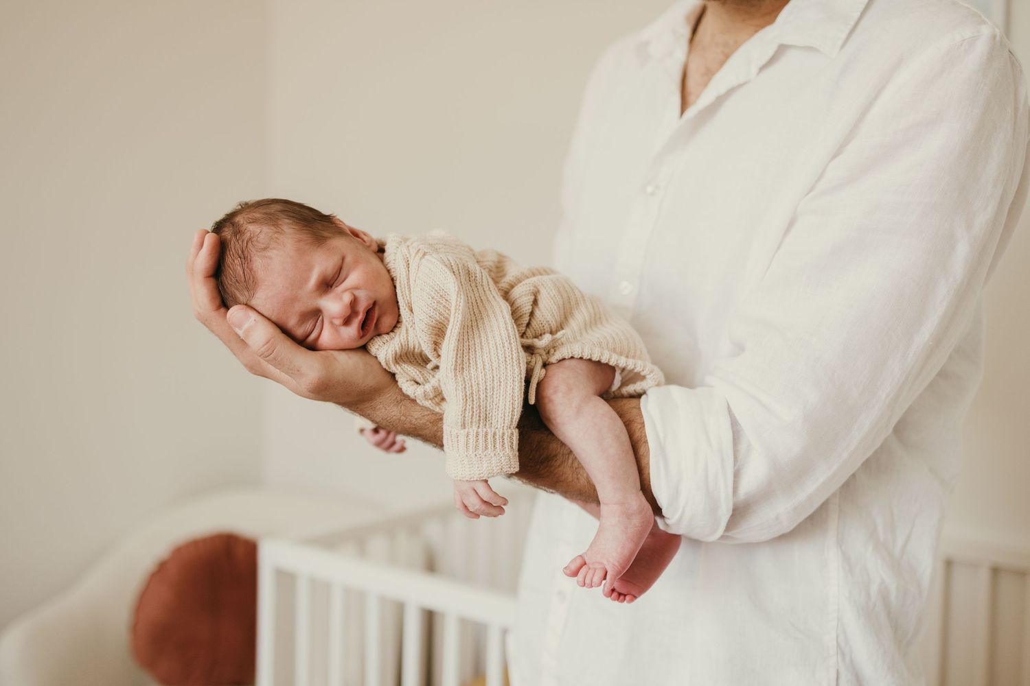 Sleeping newborn in cream knit sweater being cradled by someone in white against neutral background.