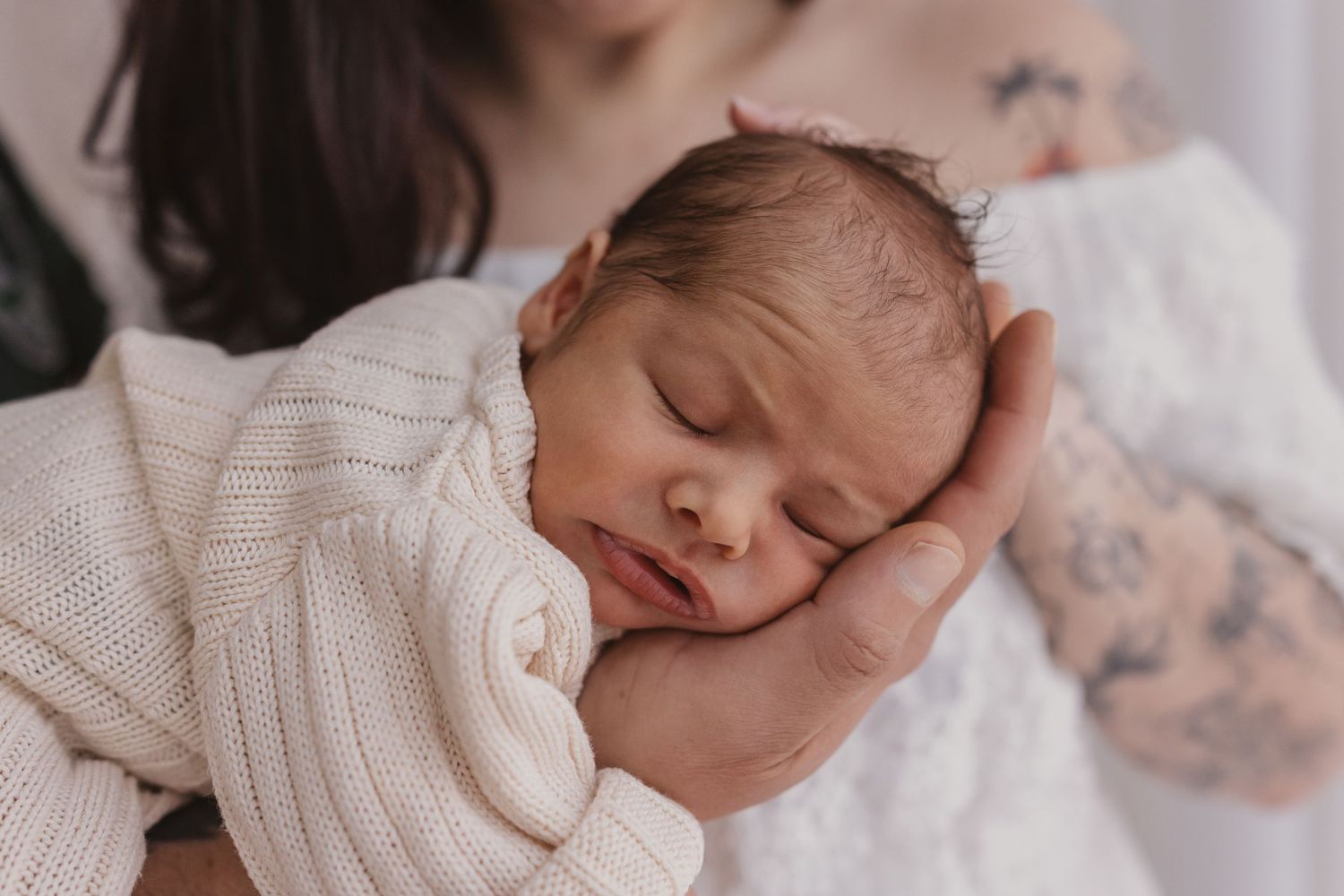Newborn baby sleeping peacefully wrapped in a cream knit blanket, cradled gently in tattooed parents hands.