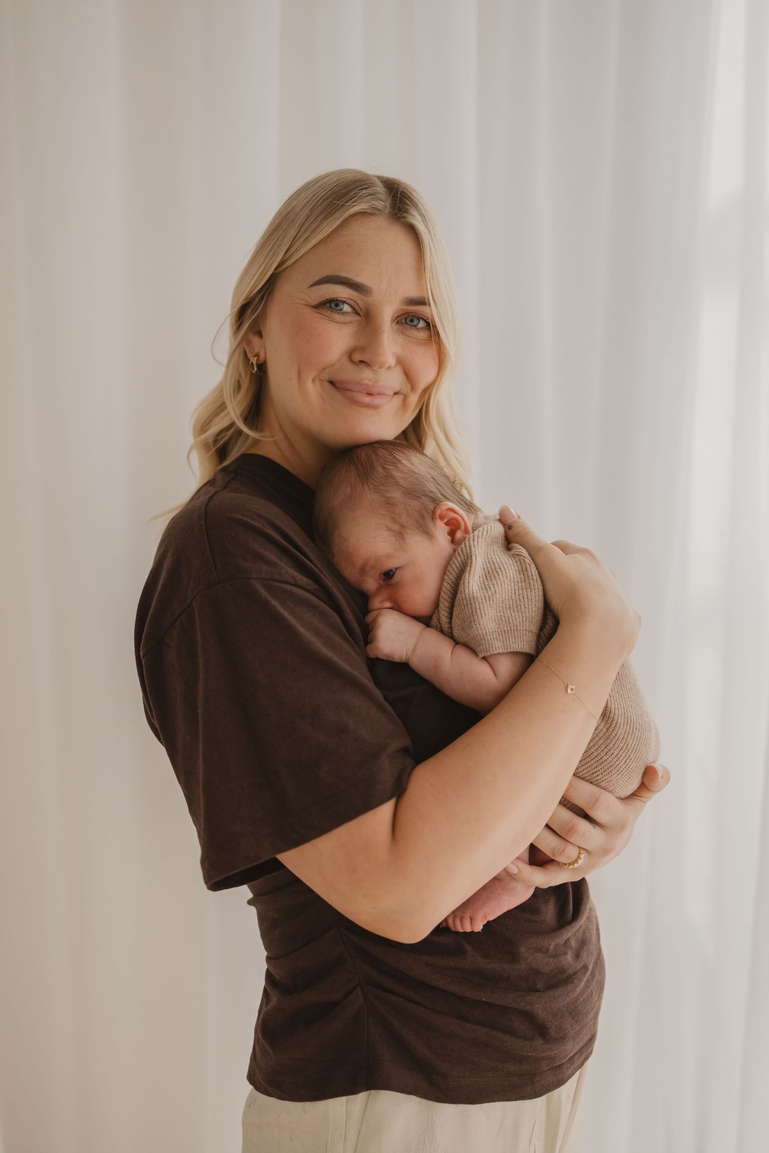 Smiling blonde woman in brown t-shirt tenderly holding a sleeping newborn baby against her chest.