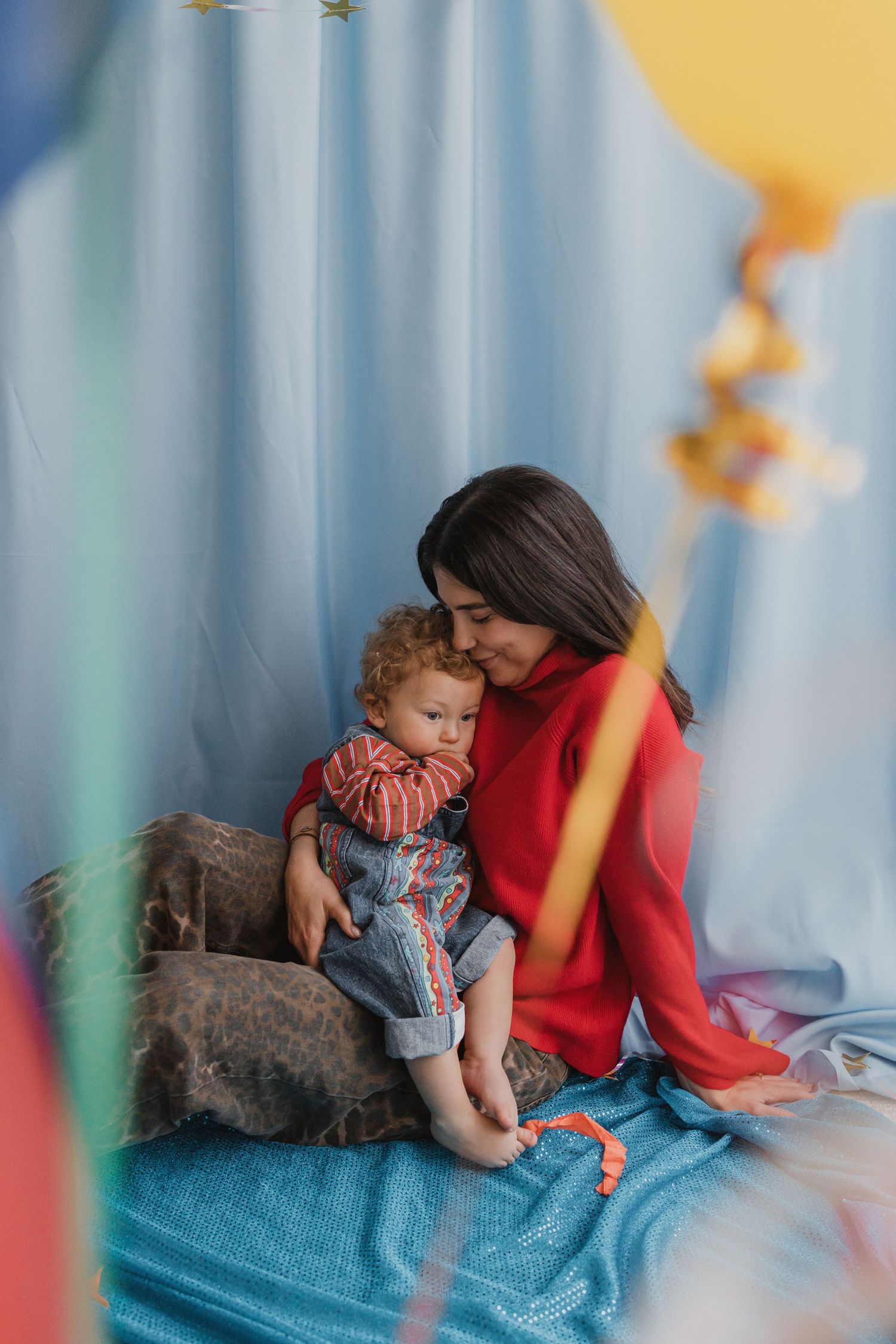 A mother in a red sweater tenderly holds her toddler in denim overalls, surrounded by colorful balloons.