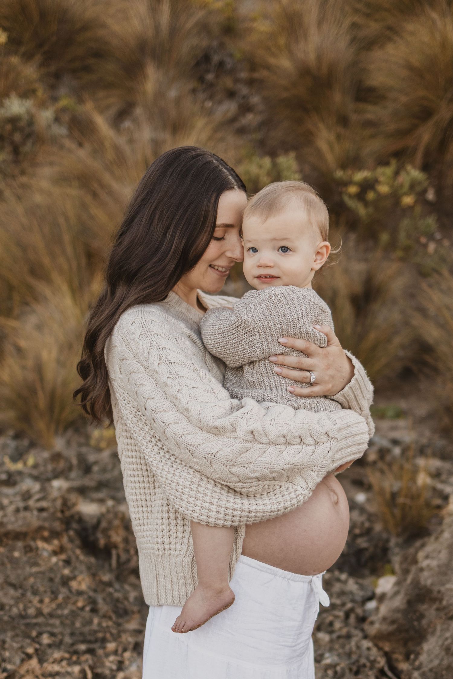 Pregnant woman in cream cable-knit sweater holding a smiling baby outdoors among golden grasses.