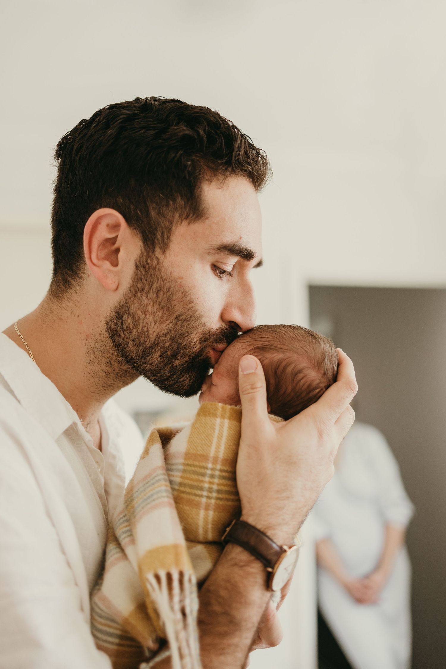 Parent in white shirt tenderly holding and kissing a newborn wrapped in plaid.