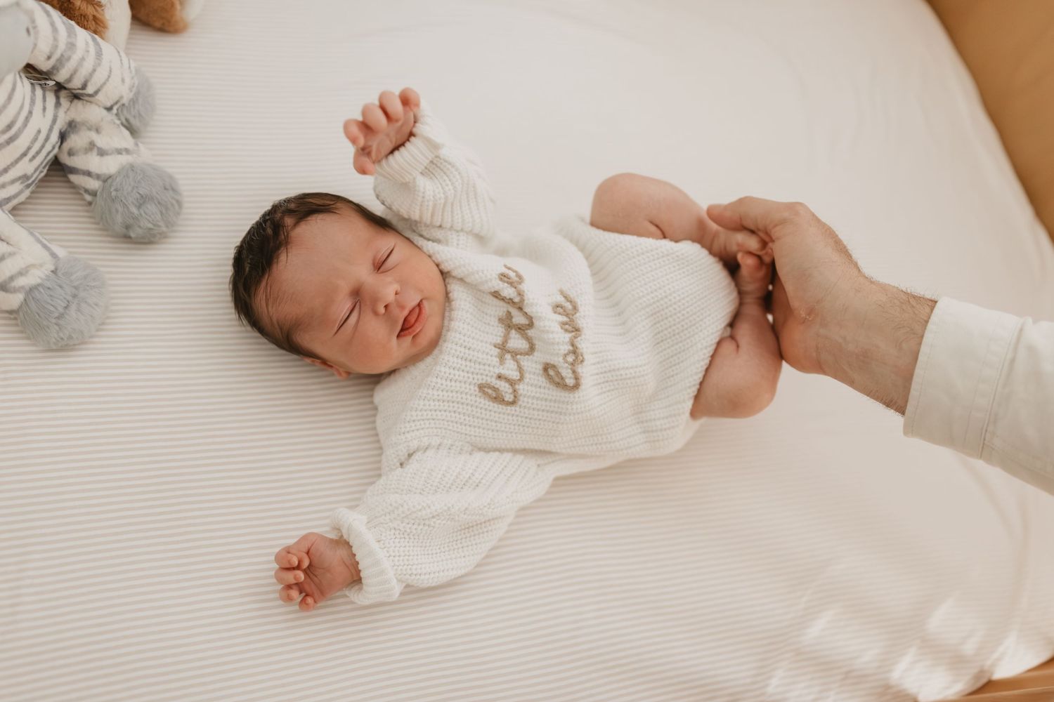 Newborn baby sleeping peacefully in white knit onesie on cream bedding, with parent's hand gently holding tiny feet.