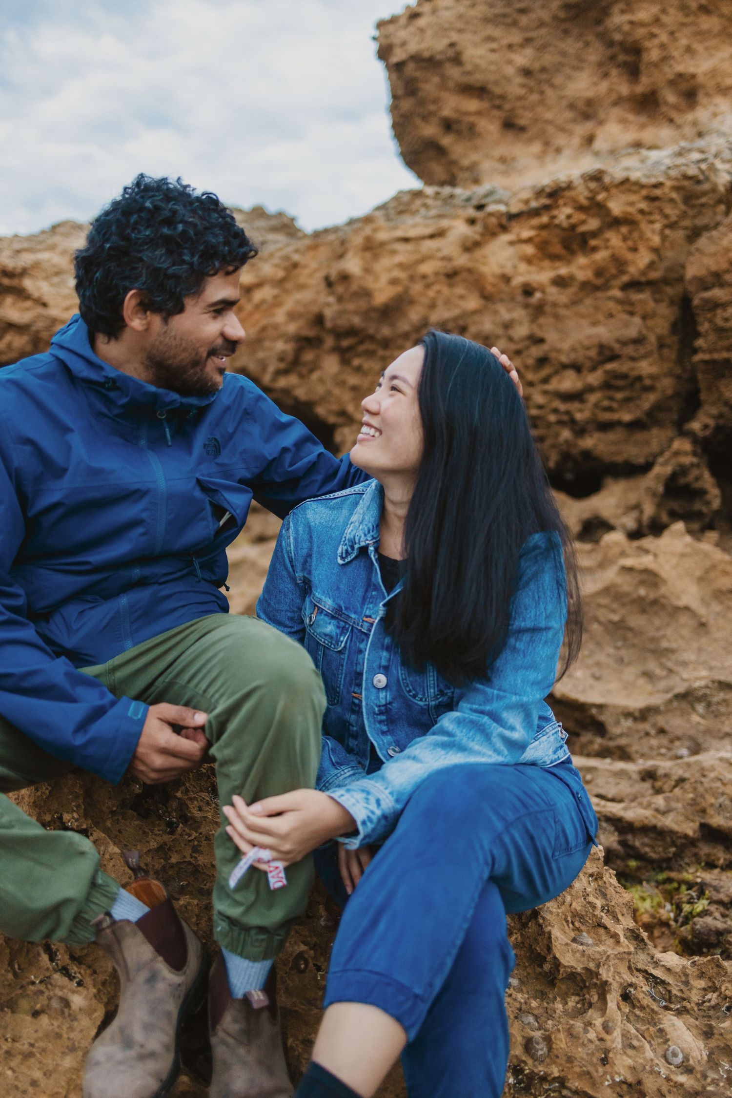 A couple sitting on rocky terrain, smiling at each other, wearing blue outfits outdoors.