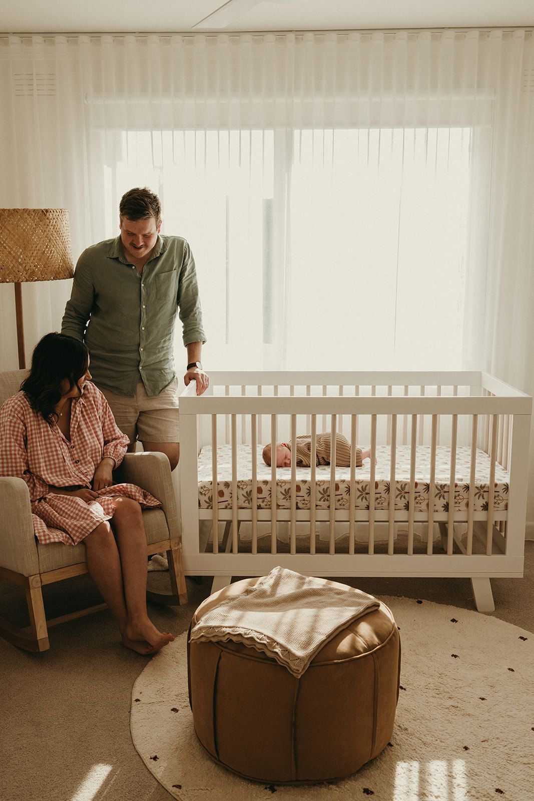 Parents admire their newborn baby resting in a white crib in a warm, neutral-toned nursery with soft natural light.