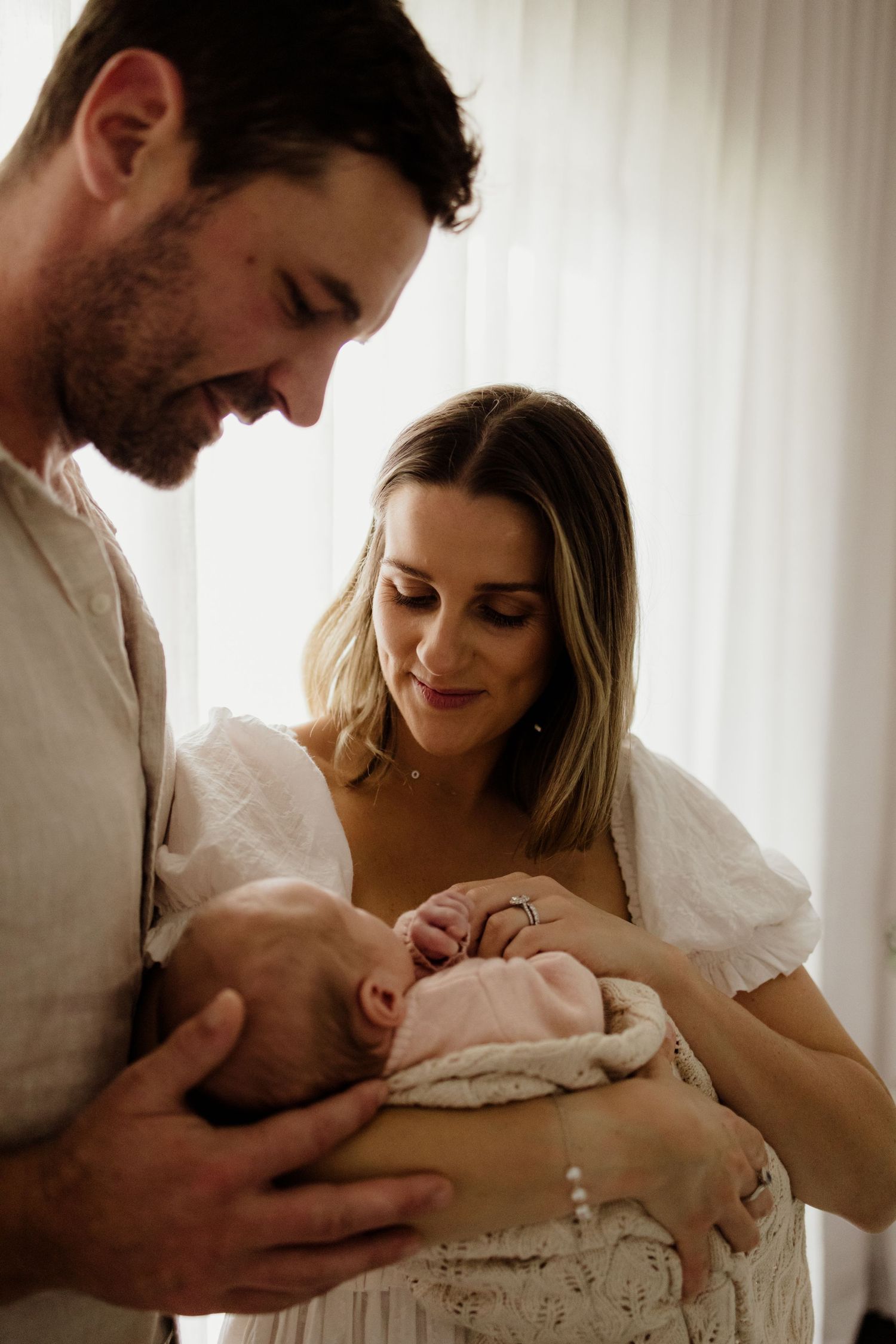 A couple lovingly gazes at their newborn baby wrapped in a pink blanket, sharing a tender family moment.