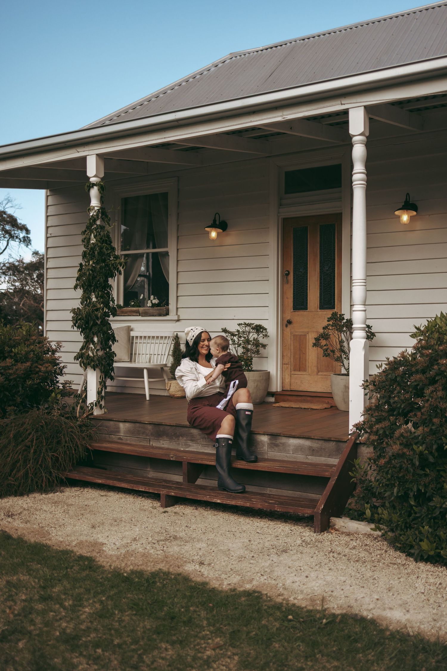 Woman sitting on porch steps of charming cottage home holding a child, with warm lantern lights and white columns.