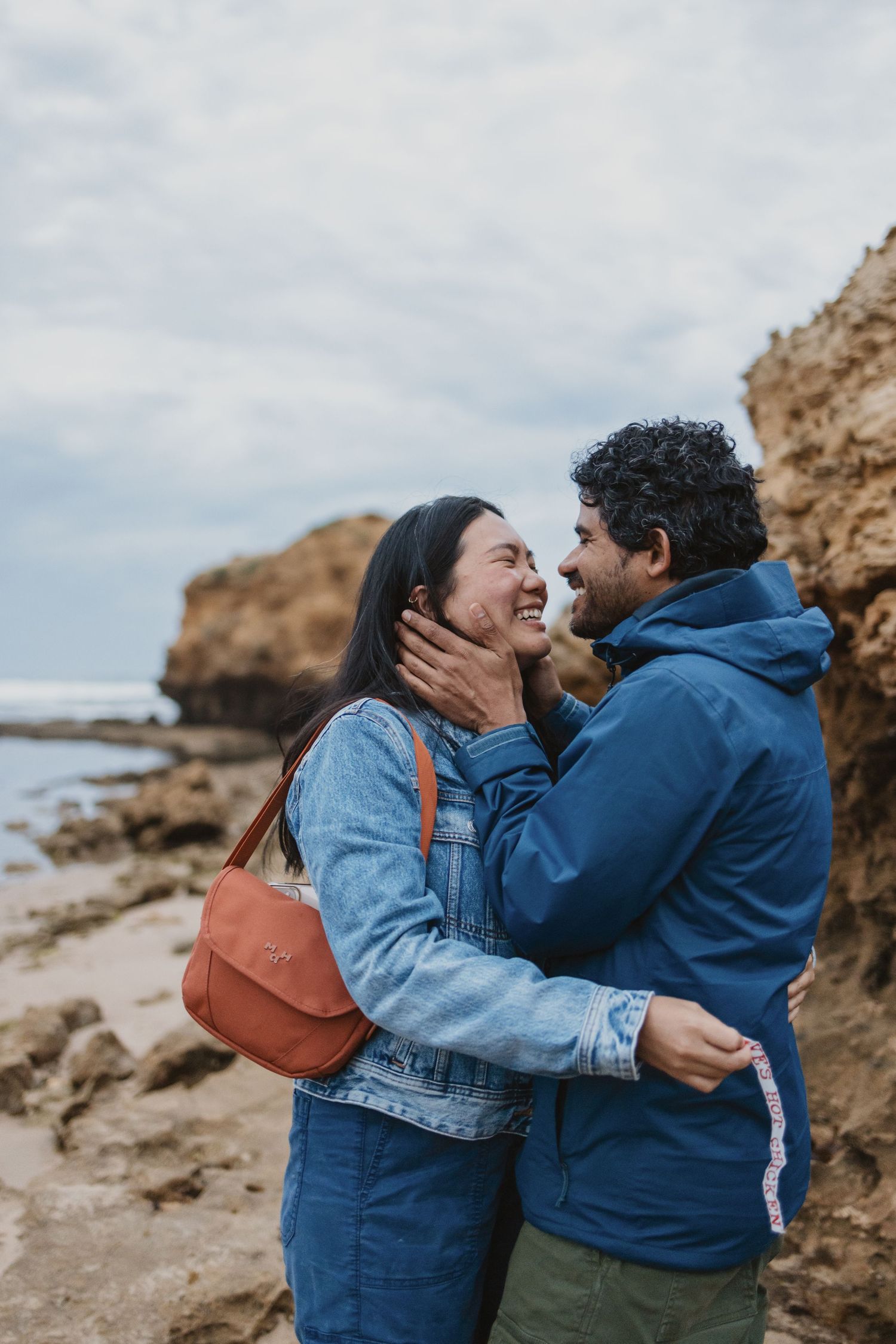 A couple laughing and embracing on a rocky beach, the man tenderly holding the woman's face as they share a joyful moment.