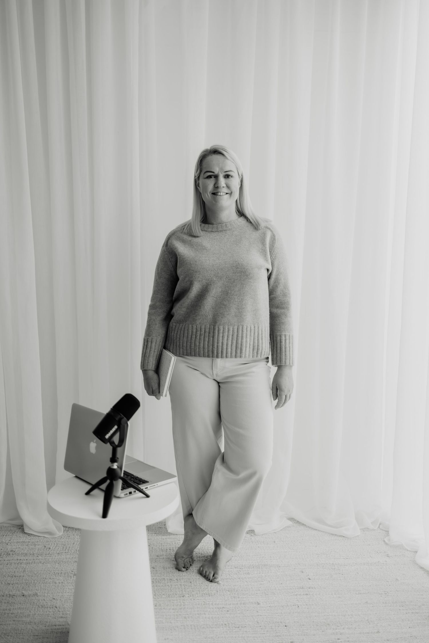 Woman in knit sweater and wide-leg pants smiles beside a microphone on a white pedestal table.