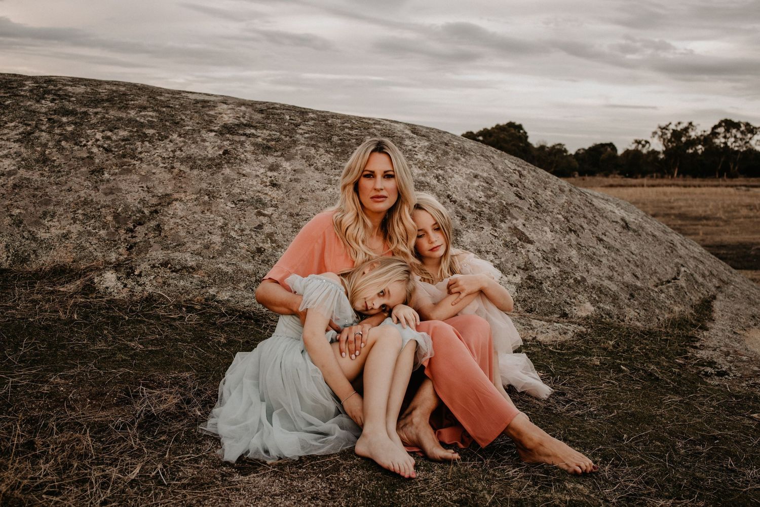 A mother sits with her two young children on a rocky outcrop in a field, wearing soft pink and white dresses at dusk.